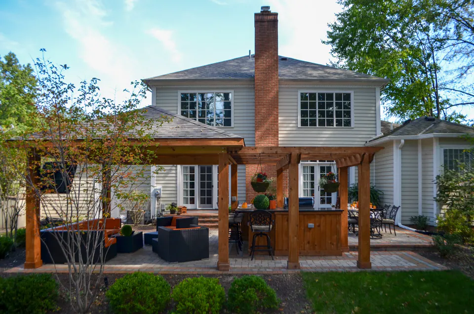 Back elevation of house with covered pavilion, outdoor kitchen, lounge seating, brick chimney, lawn, and planting.
